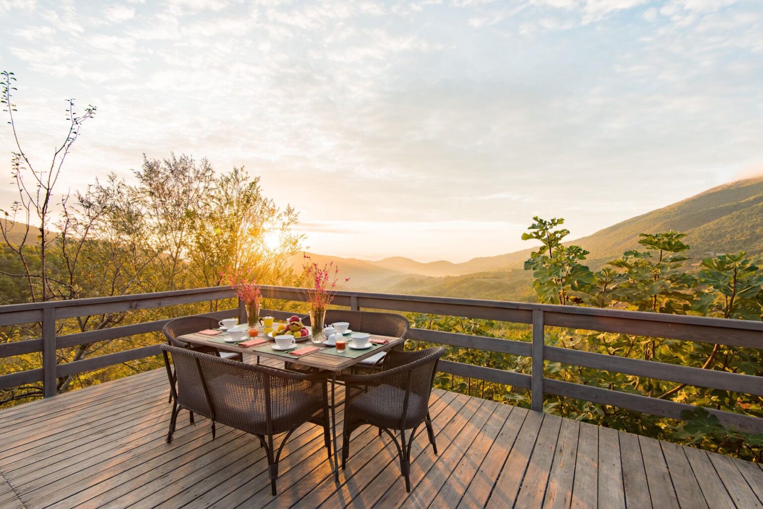 Scenic outdoor dining area with a wooden deck overlooking rolling hills at sunrise, featuring a table set with breakfast items, flowers, and chairs. | Only on SopranoVillas
