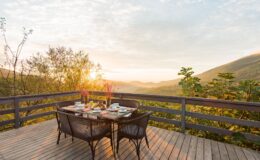 Scenic outdoor dining area with a wooden deck overlooking rolling hills at sunrise, featuring a table set with breakfast items, flowers, and chairs.