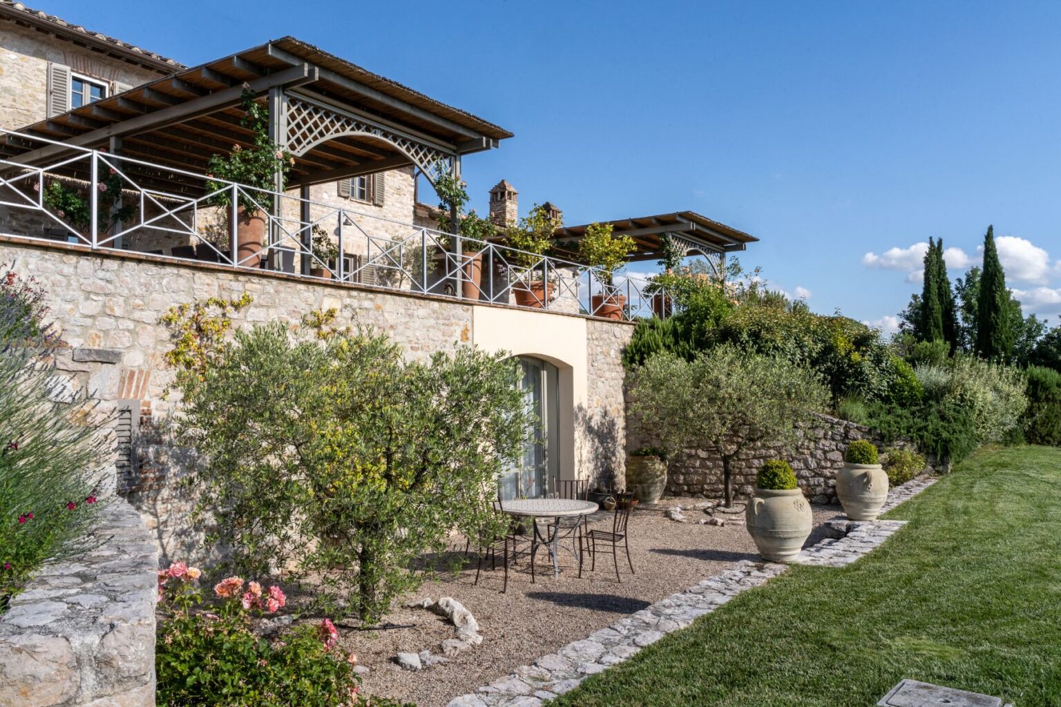 Charming stone villa with a spacious terrace and lush garden, featuring a small table and chairs surrounded by flowering plants and olive trees under a clear blue sky. | Only on SopranoVillas