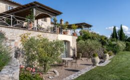 Charming stone villa with a spacious terrace and lush garden, featuring a small table and chairs surrounded by flowering plants and olive trees under a clear blue sky.