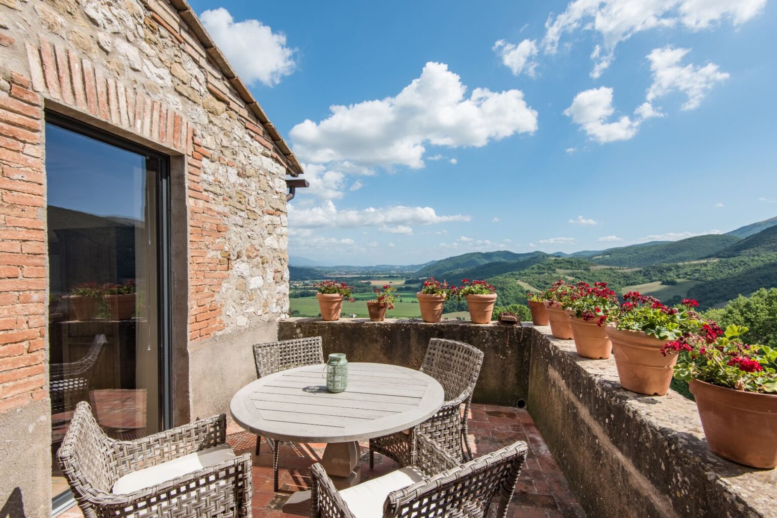 Charming terrace with a round wooden table and wicker chairs, surrounded by potted flowers, offering stunning views of rolling hills and a clear blue sky. Ideal for outdoor dining and relaxation in a picturesque setting. | Only on SopranoVillas