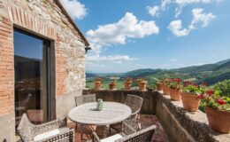 Charming terrace with a round wooden table and wicker chairs, surrounded by potted flowers, offering stunning views of rolling hills and a clear blue sky. Ideal for outdoor dining and relaxation in a picturesque setting.