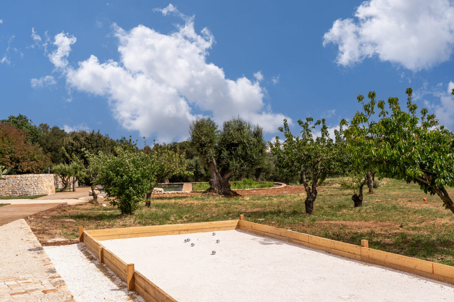 Outdoor pétanque court surrounded by lush greenery and olive trees under a blue sky with fluffy clouds, showcasing a peaceful recreational space. | Only on SopranoVillas