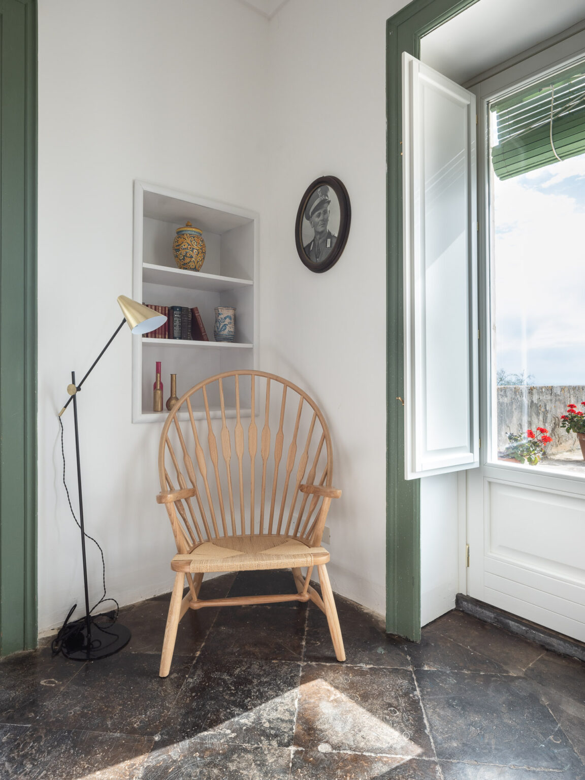Cozy reading nook featuring a wooden spindle chair, a floor lamp, and a decorative shelf with books and a vase. Natural light filters in through an open window with green blinds, illuminating the space with a warm ambiance. | Only on SopranoVillas