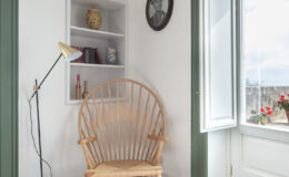Cozy reading nook featuring a wooden spindle chair, a floor lamp, and a decorative shelf with books and a vase. Natural light filters in through an open window with green blinds, illuminating the space with a warm ambiance.