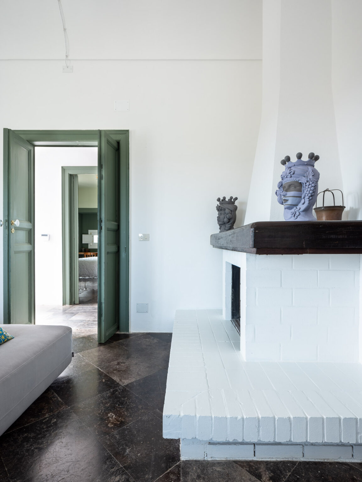 Interior view of a stylish living room featuring a white brick fireplace, a modern gray sofa, and green double doors leading to another room. Decorative ceramic sculptures adorn the mantel, and the floor showcases a unique dark patterned design, enhancing the contemporary aesthetic. | Only on SopranoVillas