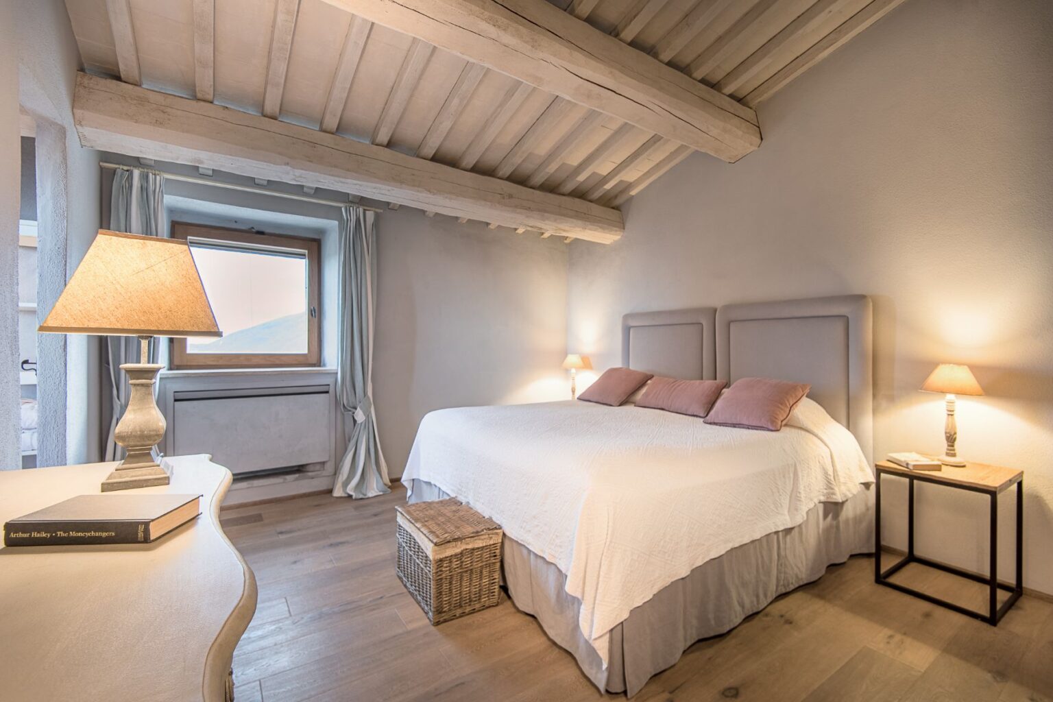 Cozy and stylish bedroom featuring a large double bed with soft pillows, a wooden ceiling with exposed beams, and warm lighting from bedside lamps. The room includes a wooden nightstand and a decorative basket, with a window allowing natural light and a view. Ideal for relaxation and comfort in a modern home setting. | Only on SopranoVillas
