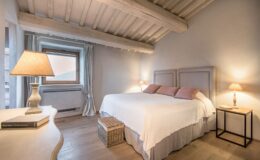 Cozy and stylish bedroom featuring a large double bed with soft pillows, a wooden ceiling with exposed beams, and warm lighting from bedside lamps. The room includes a wooden nightstand and a decorative basket, with a window allowing natural light and a view. Ideal for relaxation and comfort in a modern home setting.