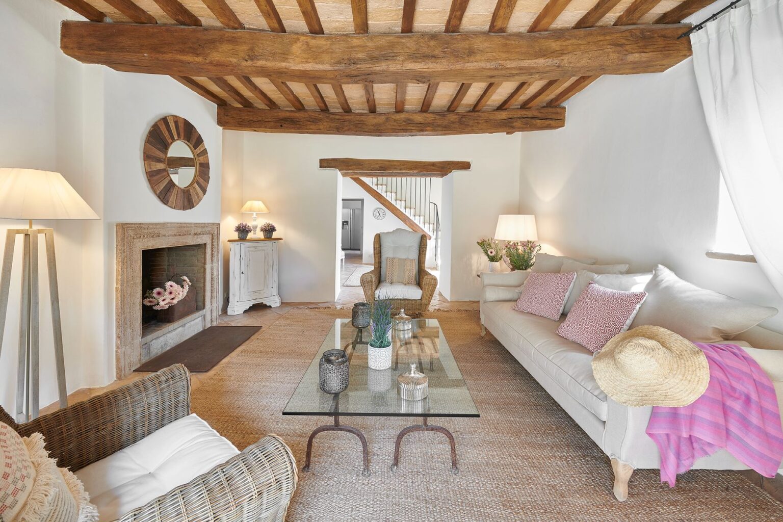 Cozy living room featuring rustic wooden beams, a stylish glass coffee table, and comfortable seating with decorative cushions. The space is illuminated by soft lamps and adorned with fresh flowers, creating a warm and inviting atmosphere. A staircase leads to an upper level, enhancing the room's charm. | Only on SopranoVillas