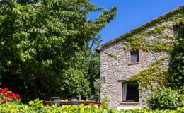 Charming stone house surrounded by lush greenery and vibrant flowers, featuring a sunny patio with outdoor seating and a clear blue sky. Ideal for a serene getaway or outdoor dining experience.