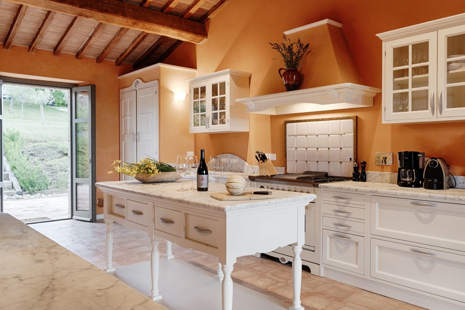 Bright and inviting kitchen featuring white cabinetry, a marble island, and rustic wooden beams. The space includes modern appliances, a wine bottle, glasses, and a decorative bowl with flowers, enhancing the warm orange walls and natural light from the open door leading to an outdoor view. | Only on SopranoVillas