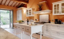 Bright and inviting kitchen featuring white cabinetry, a marble island, and rustic wooden beams. The space includes modern appliances, a wine bottle, glasses, and a decorative bowl with flowers, enhancing the warm orange walls and natural light from the open door leading to an outdoor view.