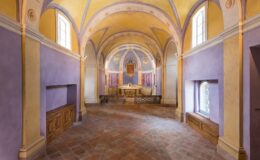 Interior view of a beautifully decorated chapel featuring arched ceilings, purple and yellow walls, and a wooden altar. The space is illuminated by natural light from windows, highlighting the intricate architectural details and serene atmosphere.