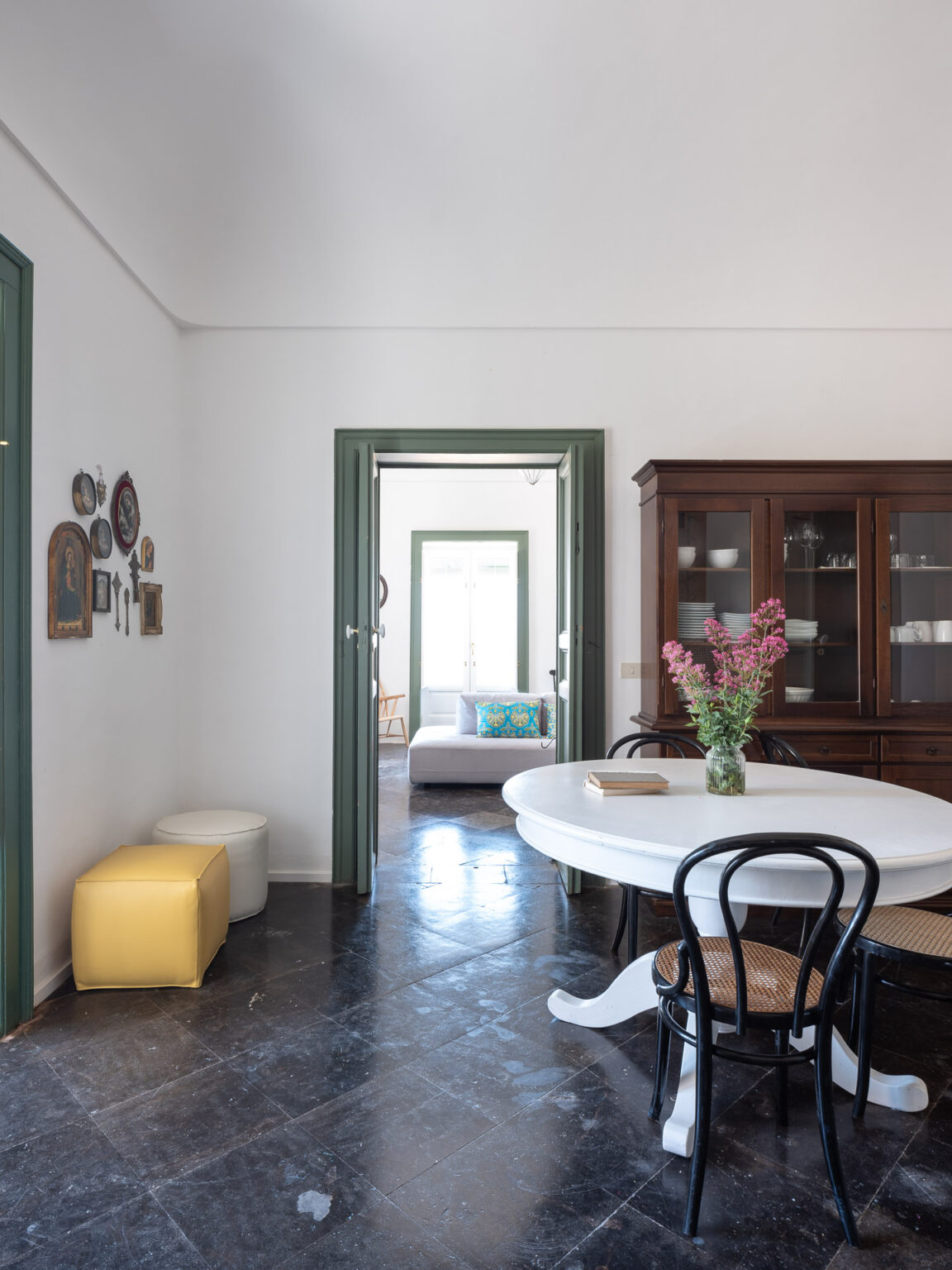 Bright and airy dining area featuring a round white table with black chairs, a wooden cabinet, and colorful decor. The space includes a view into a cozy living area, accentuated by natural light and stylish furnishings, with a yellow and white ottoman nearby. | Only on SopranoVillas