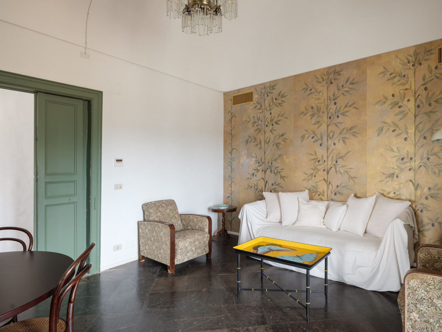 Cozy living room featuring a white sofa with decorative pillows, a colorful coffee table, and patterned armchair. The walls are adorned with elegant floral murals, complemented by a vintage chandelier and green door, creating a warm and inviting atmosphere. | Only on SopranoVillas