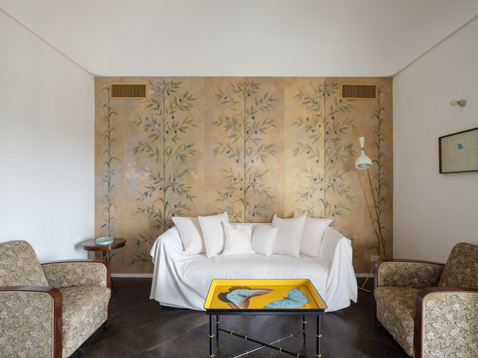 Cozy living room featuring a white sofa with plush pillows, vintage floral-patterned armchairs, and a decorative yellow coffee table with butterfly designs. The backdrop showcases elegant hand-painted botanical wall art, enhancing the room's aesthetic charm. | Only on SopranoVillas