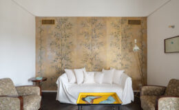 Cozy living room featuring a white sofa with plush pillows, vintage floral-patterned armchairs, and a decorative yellow coffee table with butterfly designs. The backdrop showcases elegant hand-painted botanical wall art, enhancing the room's aesthetic charm.