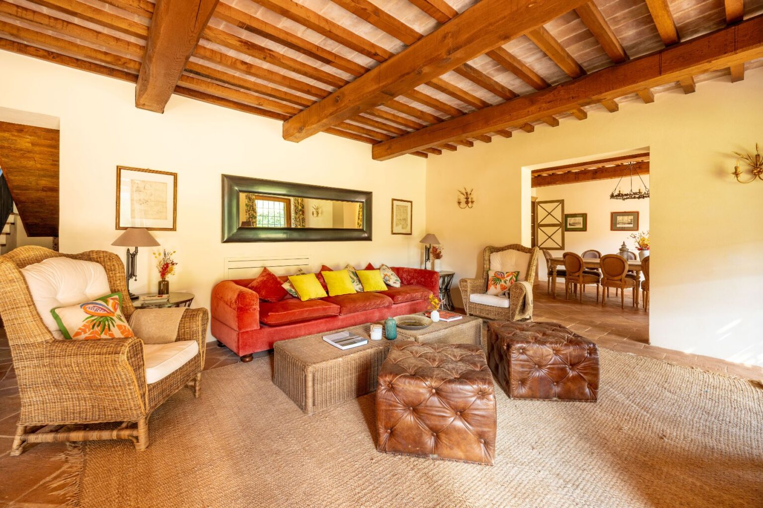 Cozy living room featuring a red sofa with colorful cushions, a woven armchair, and leather ottomans, complemented by wooden beams and natural light. A dining area is visible in the background, enhancing the warm and inviting atmosphere. Ideal for relaxation and social gatherings. | Only on SopranoVillas