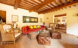 Cozy living room featuring a red sofa with colorful cushions, a woven armchair, and leather ottomans, complemented by wooden beams and natural light. A dining area is visible in the background, enhancing the warm and inviting atmosphere. Ideal for relaxation and social gatherings.