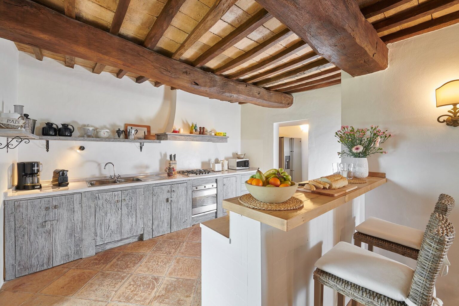 Bright and inviting kitchen featuring rustic wooden beams and cabinetry, a modern stove, and a countertop with fresh fruit and baked goods. The space is designed with a warm color palette and practical appliances, perfect for cooking and entertaining. | Only on SopranoVillas