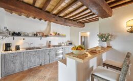 Bright and inviting kitchen featuring rustic wooden beams and cabinetry, a modern stove, and a countertop with fresh fruit and baked goods. The space is designed with a warm color palette and practical appliances, perfect for cooking and entertaining.