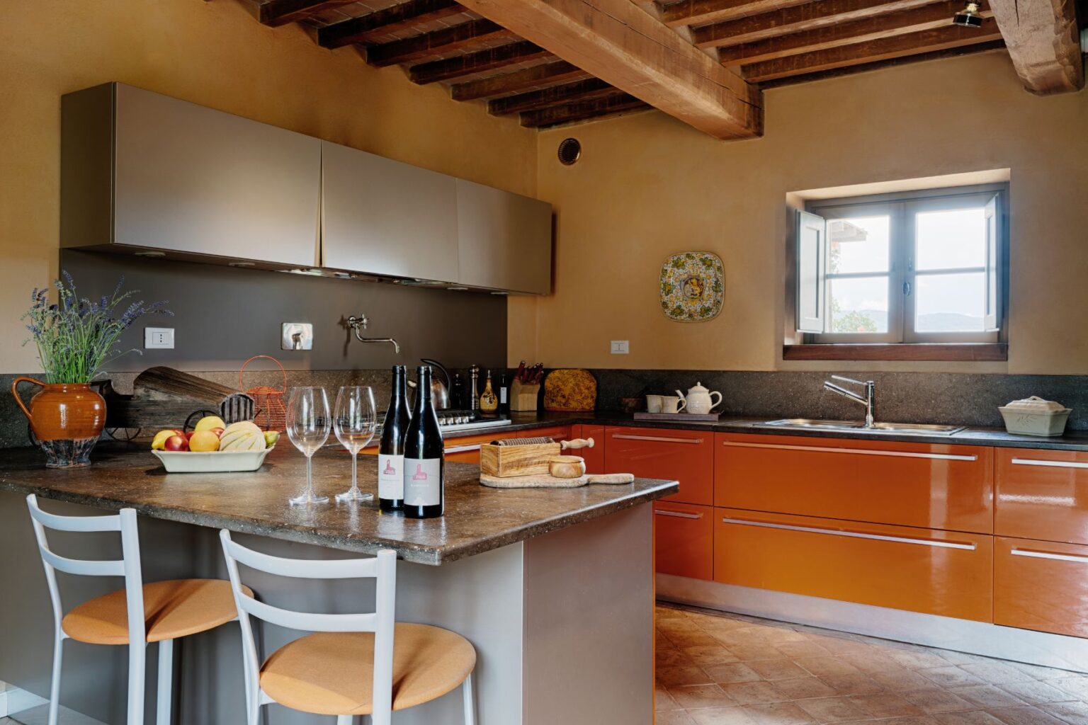 Modern kitchen interior featuring sleek orange cabinets, a gray countertop, and wooden beams. The space includes a fruit bowl, wine bottles, and elegant glassware, creating a warm and inviting atmosphere. A window offers natural light and a view of the outdoors, enhancing the cozy ambiance. | Only on SopranoVillas