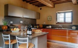 Modern kitchen interior featuring sleek orange cabinets, a gray countertop, and wooden beams. The space includes a fruit bowl, wine bottles, and elegant glassware, creating a warm and inviting atmosphere. A window offers natural light and a view of the outdoors, enhancing the cozy ambiance.