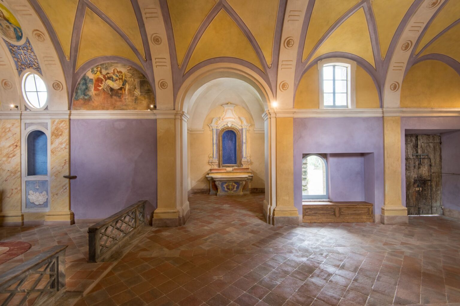 Interior view of a historic chapel featuring colorful frescoes, arched ceilings, and a central altar with a blue backdrop. The walls display a blend of yellow and purple hues, with decorative elements and wooden accents. Natural light filters through windows, enhancing the serene atmosphere of the space. | Only on SopranoVillas