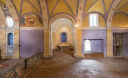 Interior view of a historic chapel featuring colorful frescoes, arched ceilings, and a central altar with a blue backdrop. The walls display a blend of yellow and purple hues, with decorative elements and wooden accents. Natural light filters through windows, enhancing the serene atmosphere of the space.