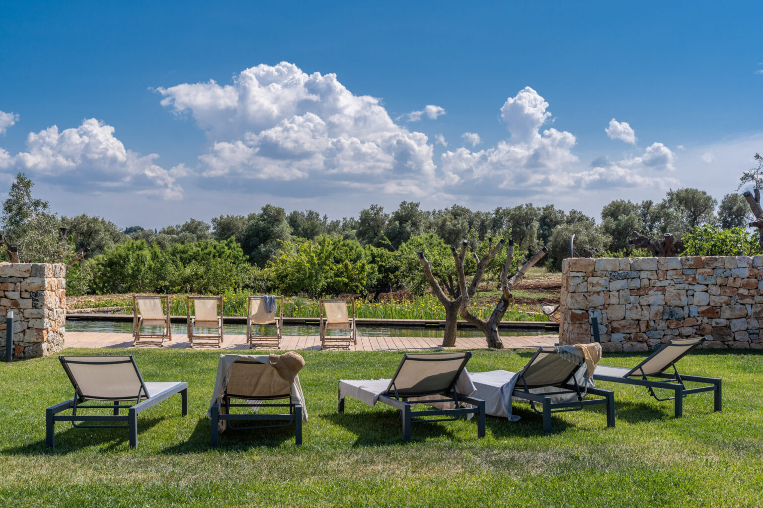 Relaxing outdoor seating area with sun loungers on a grassy lawn surrounded by trees and stone walls under a bright blue sky with fluffy clouds. Perfect for leisure and enjoying nature. | Only on SopranoVillas