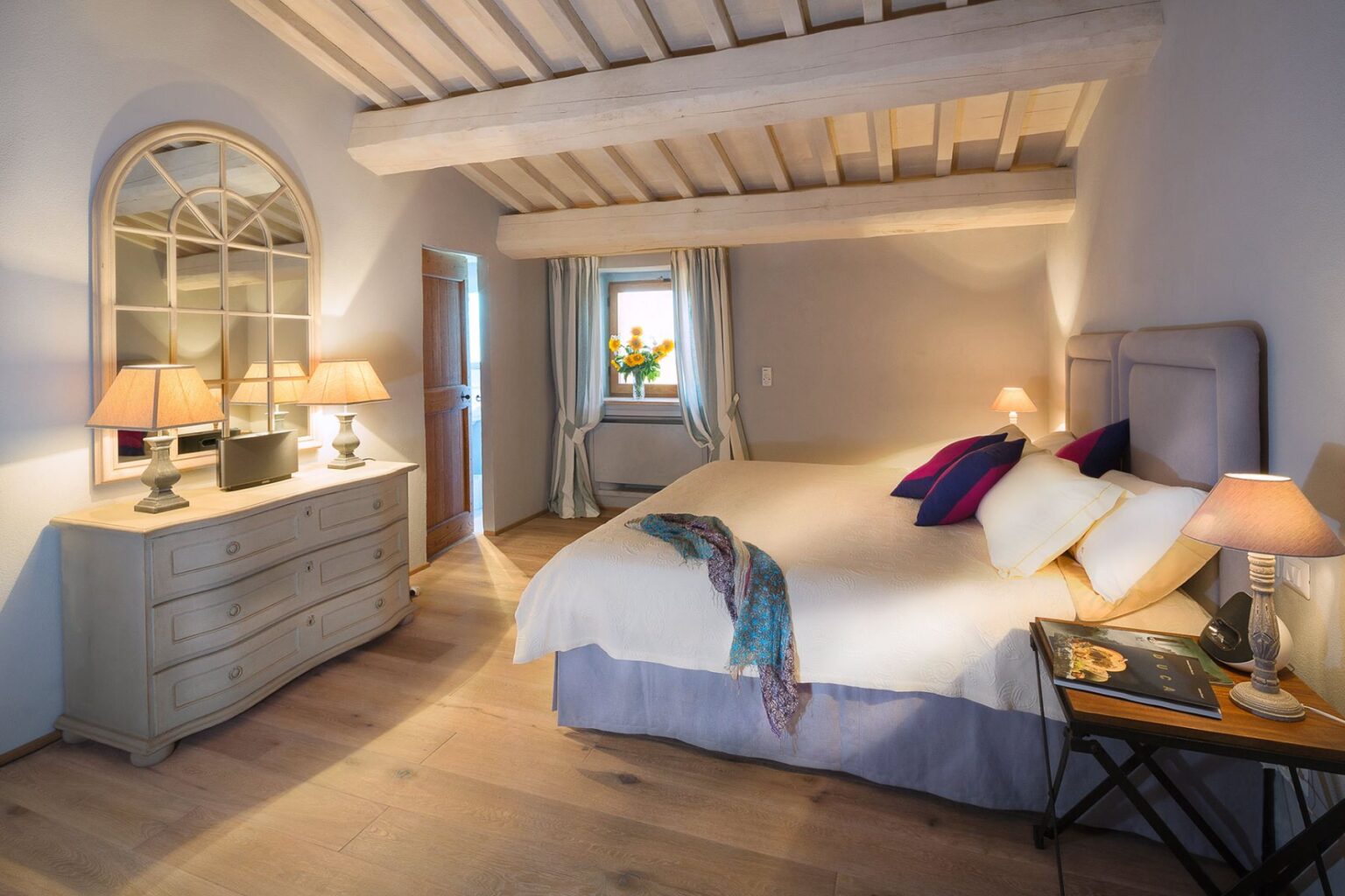 Cozy bedroom with wooden beams, soft lighting from bedside lamps, and a stylish dresser. A large bed features decorative pillows, while a window with curtains offers a view of sunflowers. The warm wood flooring enhances the inviting atmosphere. | Only on SopranoVillas