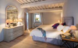 Cozy bedroom with wooden beams, soft lighting from bedside lamps, and a stylish dresser. A large bed features decorative pillows, while a window with curtains offers a view of sunflowers. The warm wood flooring enhances the inviting atmosphere.