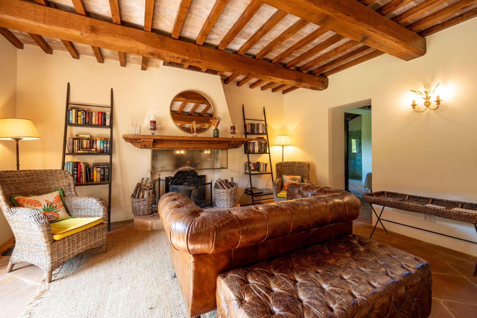 Cozy living room featuring rustic wooden beams, a leather sofa, and a woven armchair. A bookshelf filled with books and a decorative fireplace add charm, while natural light enhances the inviting atmosphere. Ideal for relaxation and reading in a warm, homey setting. | Only on SopranoVillas