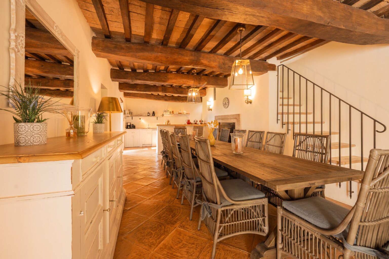 Spacious rustic dining area featuring a long wooden table surrounded by stylish wicker chairs, illuminated by pendant lights. The room showcases exposed wooden beams, terracotta tile flooring, and a warm color palette, creating a cozy atmosphere. A well-equipped kitchen is visible in the background, enhancing the inviting setting for gatherings. | Only on SopranoVillas