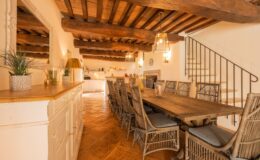 Spacious rustic dining area featuring a long wooden table surrounded by stylish wicker chairs, illuminated by pendant lights. The room showcases exposed wooden beams, terracotta tile flooring, and a warm color palette, creating a cozy atmosphere. A well-equipped kitchen is visible in the background, enhancing the inviting setting for gatherings.