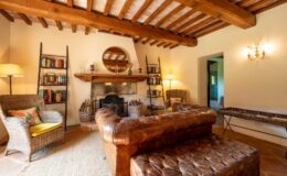 Cozy living room featuring rustic wooden beams, a leather sofa, and a woven armchair. A bookshelf filled with books and a decorative fireplace add charm, while natural light enhances the inviting atmosphere. Ideal for relaxation and reading in a warm, homey setting.