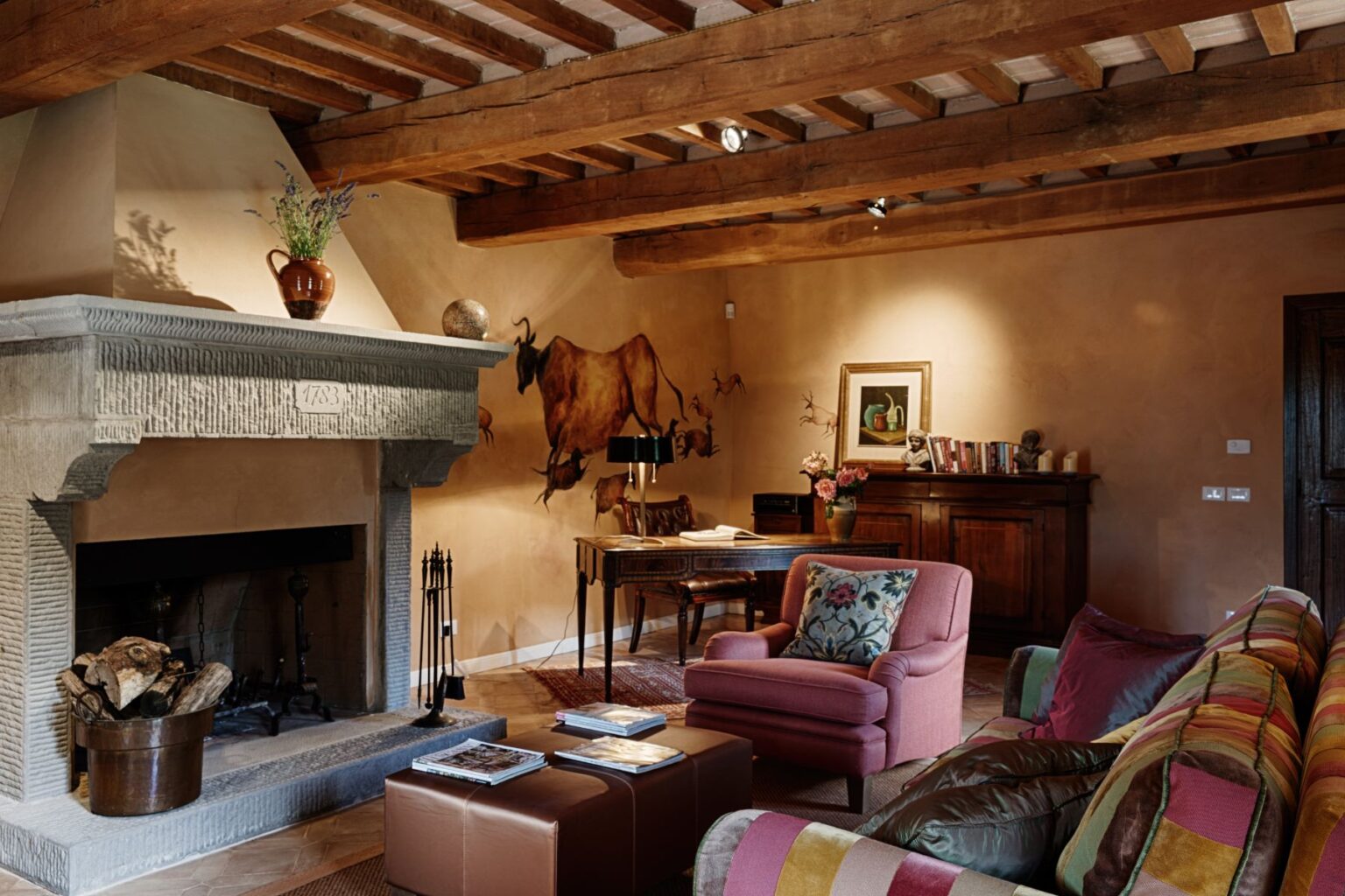 Cozy living room featuring a stone fireplace with a decorative flower vase, rustic wooden beams, and artistic wall murals. The space includes a colorful sofa, a plush armchair, a wooden desk, and a bookshelf, creating an inviting atmosphere for relaxation and conversation. | Only on SopranoVillas