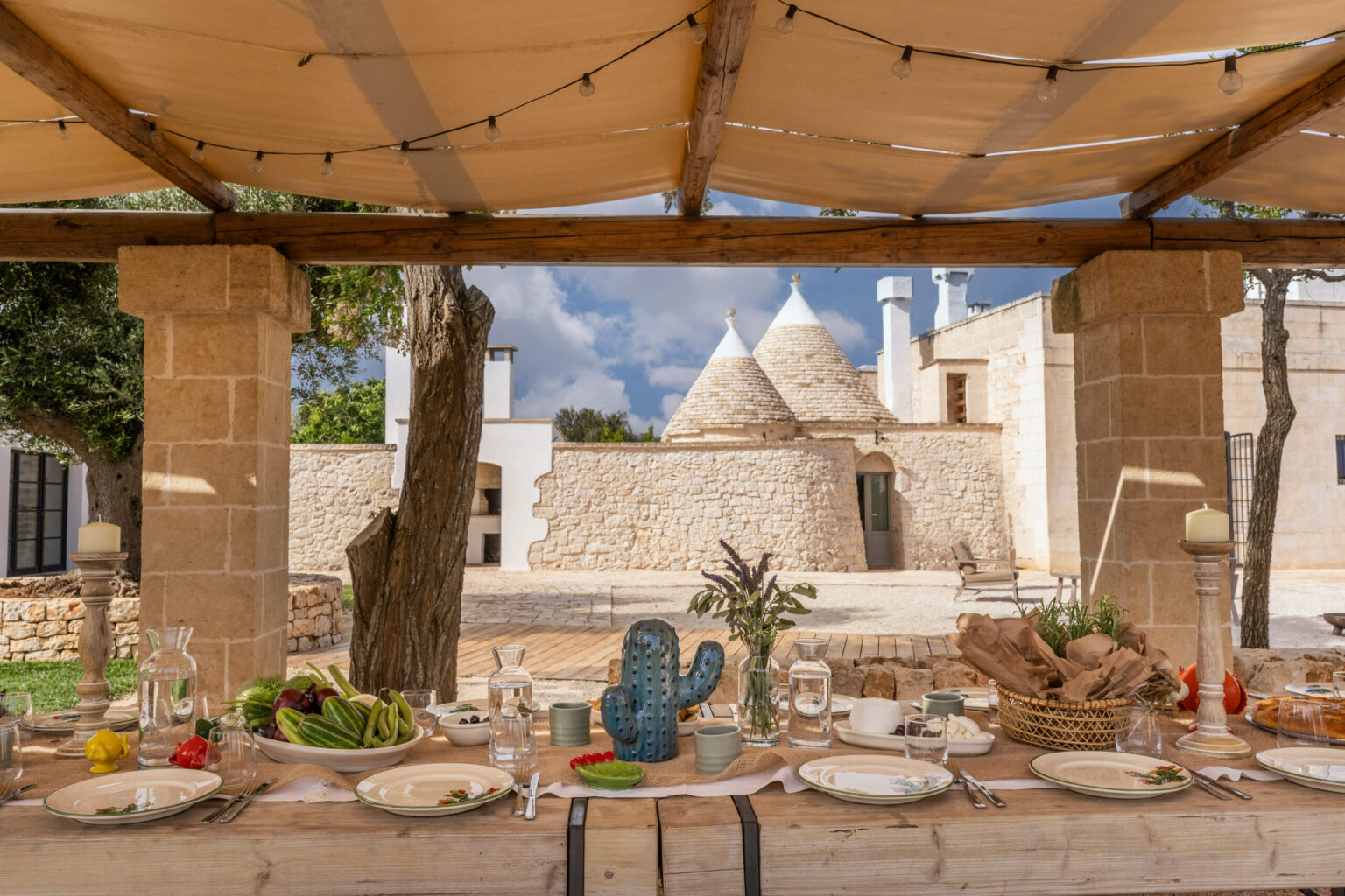 Outdoor dining setup with a beautifully arranged table featuring plates and fresh produce, set against the backdrop of a traditional trullo house in a sunny landscape, showcasing rustic charm and inviting ambiance. | Only on SopranoVillas