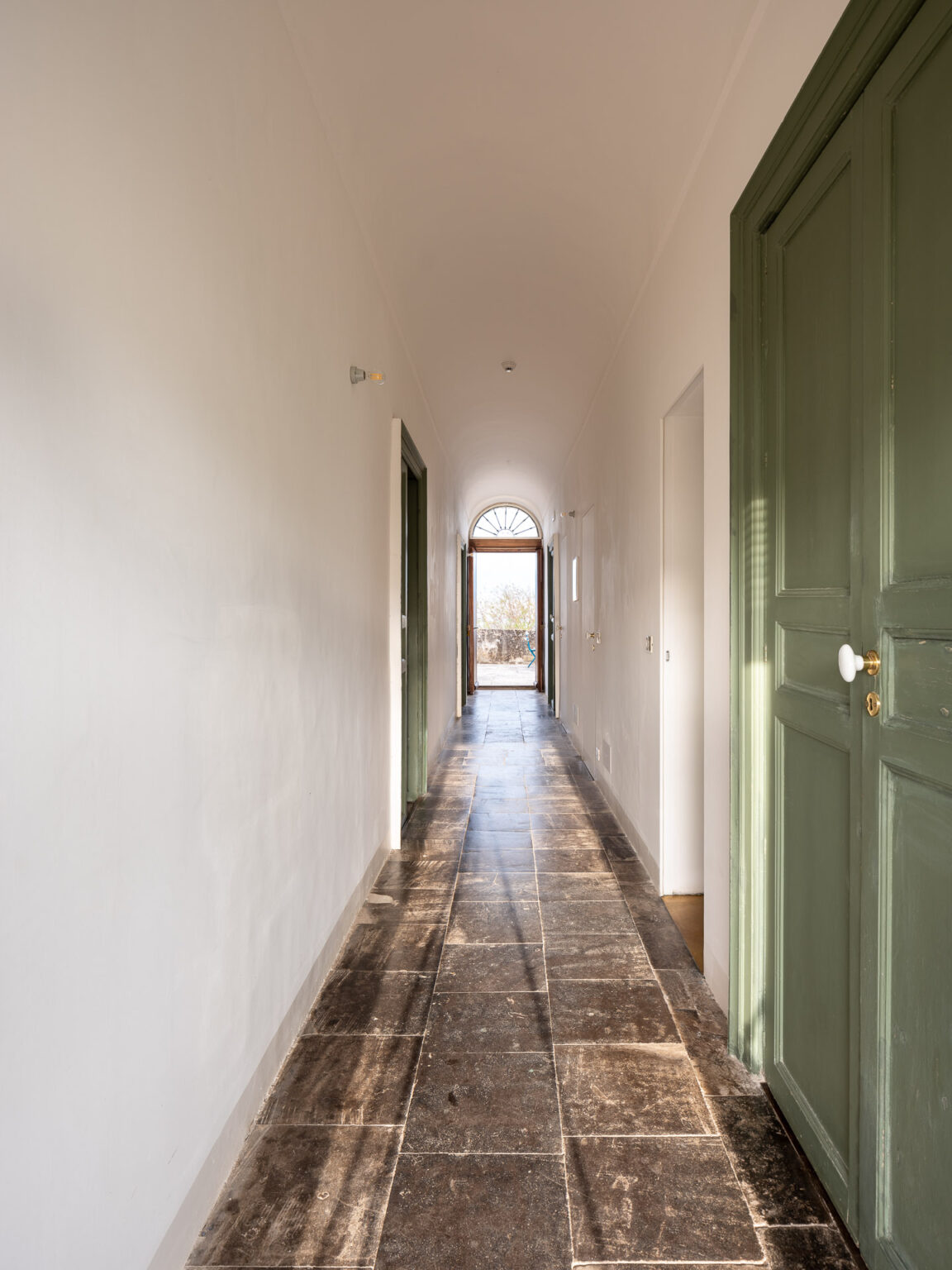 Long hallway with stone flooring and green doors, leading to an arched doorway that opens to an outdoor view, illuminated by natural light. | Only on SopranoVillas