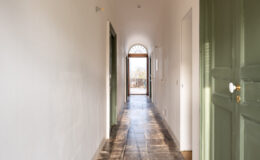 Long hallway with stone flooring and green doors, leading to an arched doorway that opens to an outdoor view, illuminated by natural light.