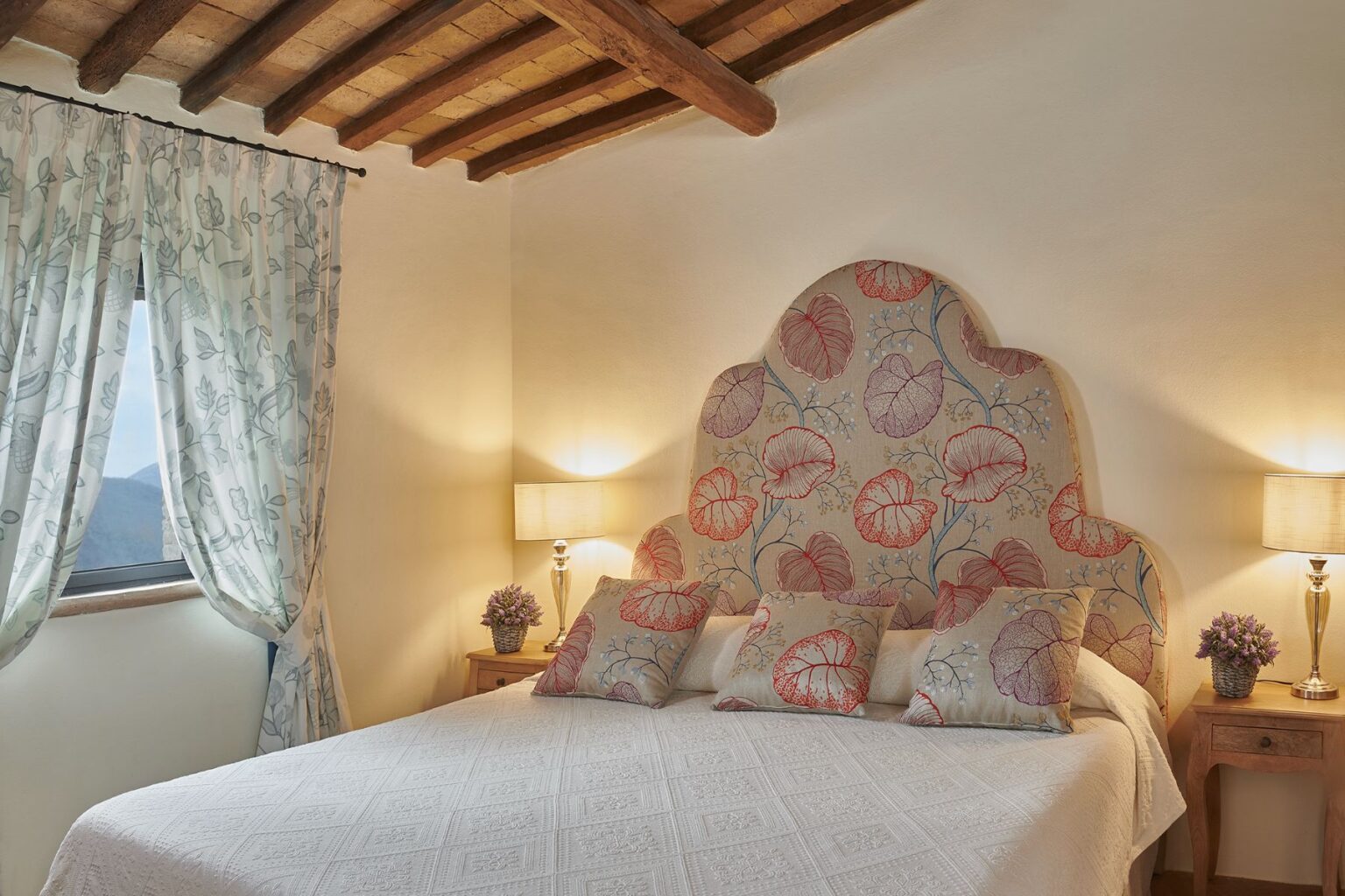 Cozy bedroom featuring a floral-patterned headboard, decorative pillows, and soft lighting. The room is adorned with light curtains, a bedside table with a vase of flowers, and exposed wooden beams on the ceiling, creating a warm and inviting atmosphere. | Only on SopranoVillas