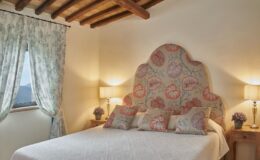 Cozy bedroom featuring a floral-patterned headboard, decorative pillows, and soft lighting. The room is adorned with light curtains, a bedside table with a vase of flowers, and exposed wooden beams on the ceiling, creating a warm and inviting atmosphere.