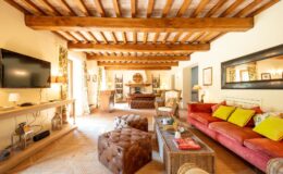 Spacious living room featuring wooden beams, a large flat-screen TV, and a cozy seating area with colorful cushions. The room includes a rustic coffee table, stylish lighting, and decorative curtains, creating a warm and inviting atmosphere.
