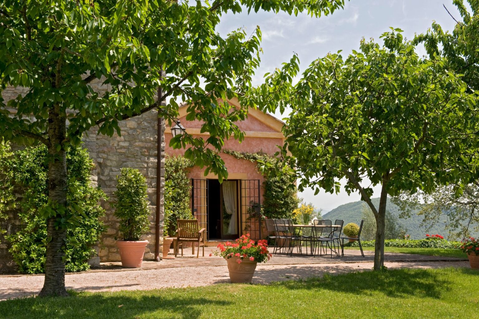 Charming countryside villa with a stone exterior, surrounded by lush greenery and blooming flowers, featuring outdoor seating and a scenic view of the mountains. Ideal for relaxation and enjoying nature. | Only on SopranoVillas