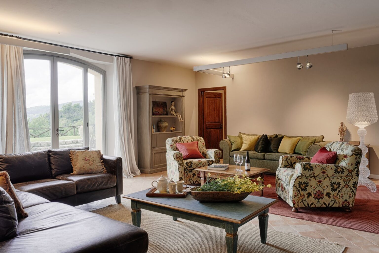 Cozy living room featuring a mix of modern and vintage furniture, including a leather sofa, floral armchairs, and a green couch with decorative pillows. A wooden coffee table holds a tray with tea service and flowers, while large windows provide natural light and scenic views. | Only on SopranoVillas
