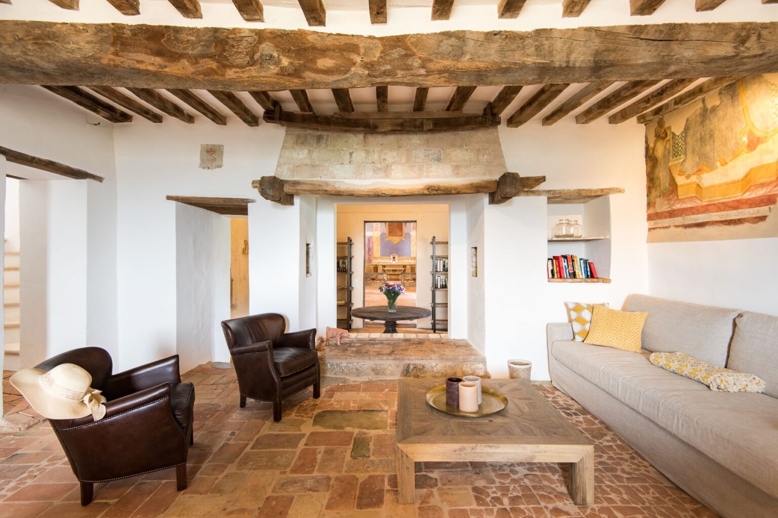 Cozy living room featuring rustic wooden beams, a stone fireplace, and terracotta flooring. The space includes two brown leather armchairs, a wooden coffee table, and a light gray sofa with decorative pillows. A small bookshelf and a colorful floral arrangement add charm to the inviting atmosphere. | Only on SopranoVillas