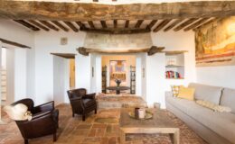 Cozy living room featuring rustic wooden beams, a stone fireplace, and terracotta flooring. The space includes two brown leather armchairs, a wooden coffee table, and a light gray sofa with decorative pillows. A small bookshelf and a colorful floral arrangement add charm to the inviting atmosphere.
