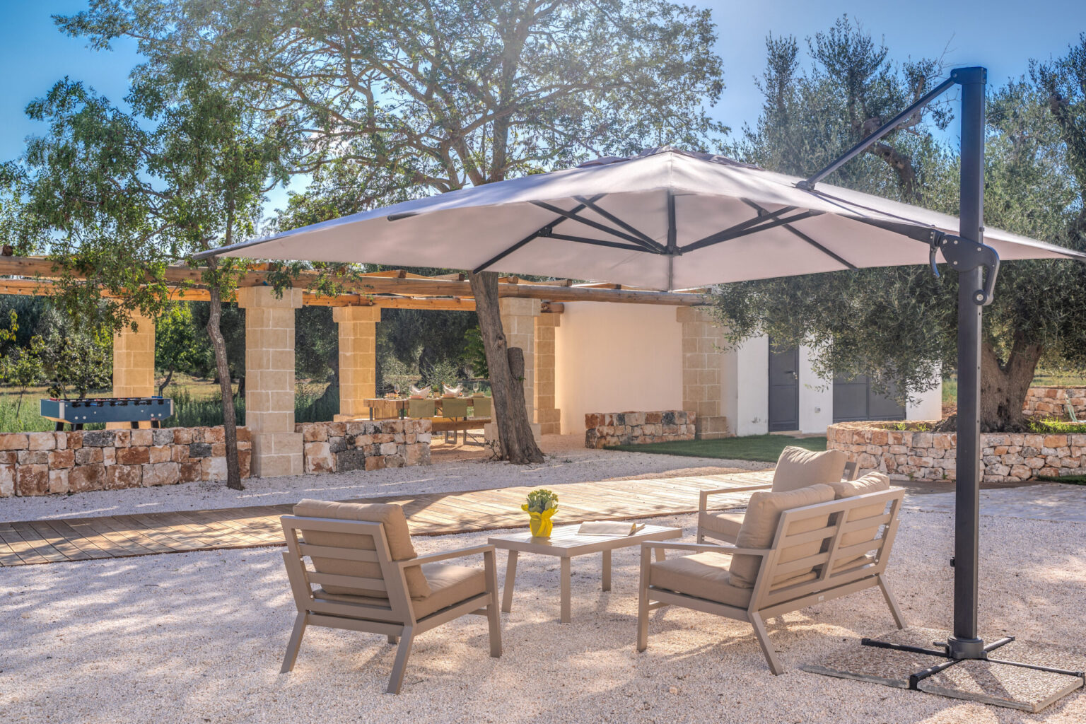 Cozy outdoor seating area featuring two wooden chairs and a small table under a large umbrella, surrounded by lush greenery and a stone wall, ideal for relaxation and entertaining. | Only on SopranoVillas