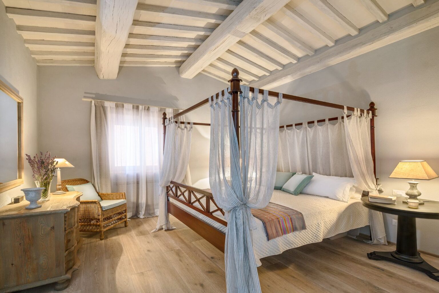 Cozy bedroom featuring a four-poster bed with sheer drapes, wooden ceiling beams, and a warm color palette. The room includes a comfortable armchair, a nightstand with a lamp, and a decorative vase with flowers, creating a serene and inviting atmosphere. | Only on SopranoVillas