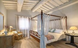Cozy bedroom featuring a four-poster bed with sheer drapes, wooden ceiling beams, and a warm color palette. The room includes a comfortable armchair, a nightstand with a lamp, and a decorative vase with flowers, creating a serene and inviting atmosphere.